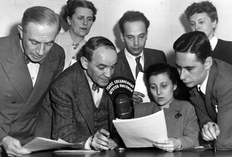 “Radio Broadcast Sent To Russia By State Department” photograph from the National Archives, Harry S. Truman Library and Museum. Description: Interior view of seven men and women taken during a radio broadcast sent to Russia from the State Department’s studios in New York. Identified as left to right: Boris Brodenov, Kathrine Elene, James Shigorin, Vladmir Postman, Mrs. Lucy Bates, Victor Franzusoff, and Mrs. Tatiana Hecker, all American citizens. Lettering on top of microphone is in Russian language. (Charles Thayer supervised the programs.) Date(s): ca. February 1947.