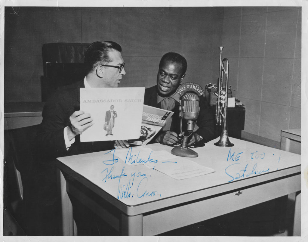 Willis Conover i Louis Armstrong siedzą przy stole w studiu Głosu Ameryki w Waszyngtonie, obok mikrofonu Voice of America i trąbki; trzymają płytę „Ambassador Satch”, 1956.