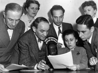 “Radio Broadcast Sent To Russia By State Department” photograph from the National Archives, Harry S. Truman Library and Museum. Description: Interior view of seven men and women taken during a radio broadcast sent to Russia from the State Department’s studios in New York. Identified as left to right: Boris Brodenov, Kathrine Elene, James Shigorin, Vladmir Postman, Mrs. Lucy Bates, Victor Franzusoff, and Mrs. Tatiana Hecker, all American citizens. Lettering on top of microphone is in Russian language. (Charles Thayer supervised the programs.) Date(s): ca. February 1947.